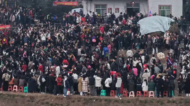 Hundreds of people can be seen standing on the streets outside a white bricked house. The picture is taken from a distance.